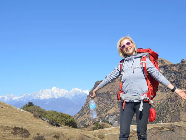 A smiling woman wearing a backpack and sunglasses standing with open arms on a grassy mountain ridge with snowy Himalayan peaks in the background