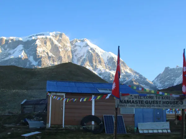 Namaste Guest House at Kori Danda in Nepal with prayer flags, national flags, and snow-capped Himalayan mountains in the background under a clear blue sky.