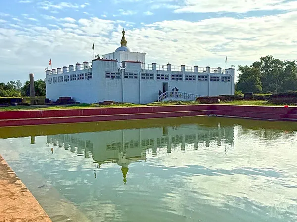 The white Maya Devi Temple building and Ashoka Pillar reflecting in the sacred Puskarini pond under a partly cloudy sky in Lumbini