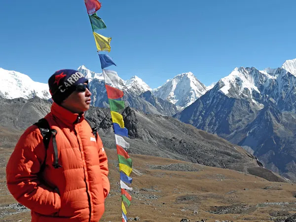Trekker wearing a red down jacket standing next to colorful prayer flags with the snow-covered Langtang mountain range behind