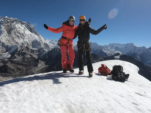 Two triumphantly celebrating mountaineers standing on a snow peak with Mount Everest rising behind them under a clear blue sky