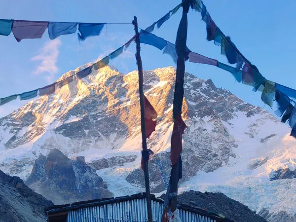 Mount Makalu's massive snow-covered peak illuminated by golden sunlight during sunrise or sunset, framed behind colorful Buddhist prayer flags strung across wooden poles near a campsite