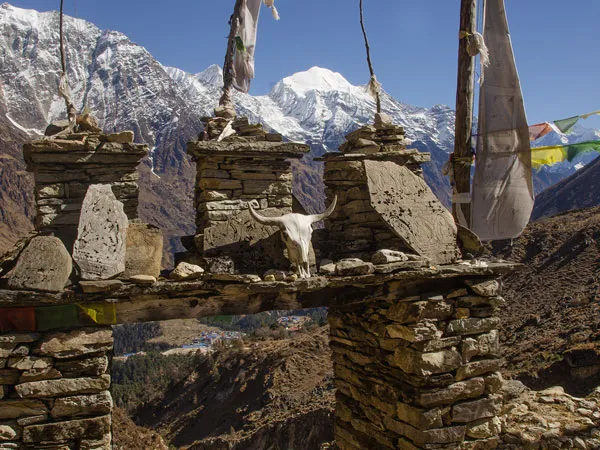 Traditional stone gateway decorated with Mani stones, prayer flags, and an animal skull against snow-capped Himalayan peaks in Tsum Valley