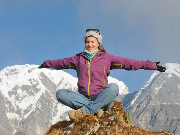 A female trekker sitting cross-legged with open arms on a mountain peak during the 10-day Mardi Himal Trek, framed by snow-capped Annapurna Himalayan mountains and a clear sky.