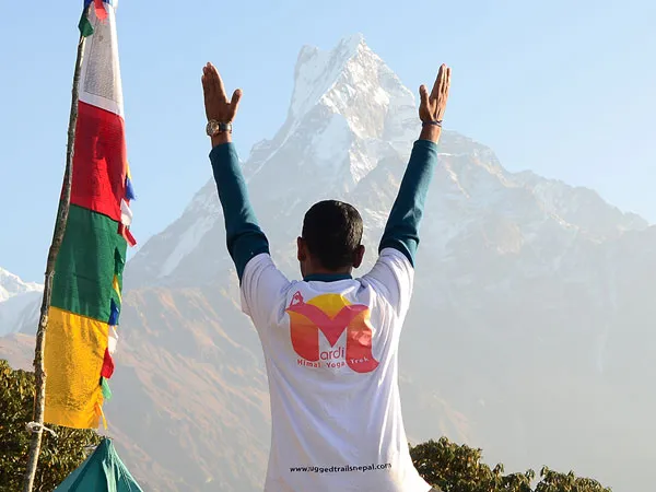 A trekker wearing a Mardi Himal Yoga Trek t-shirt stands with raised arms facing the iconic peak of Mount Machhapuchhre (Fishtail) alongside colorful prayer flags.