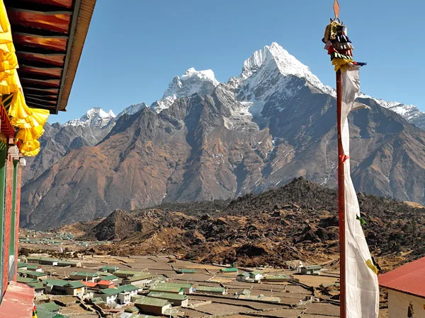 A high-altitude Sherpa village nestled in a valley beneath snow-capped Himalayan peaks, viewed from a monastery roof with a traditional white prayer pole