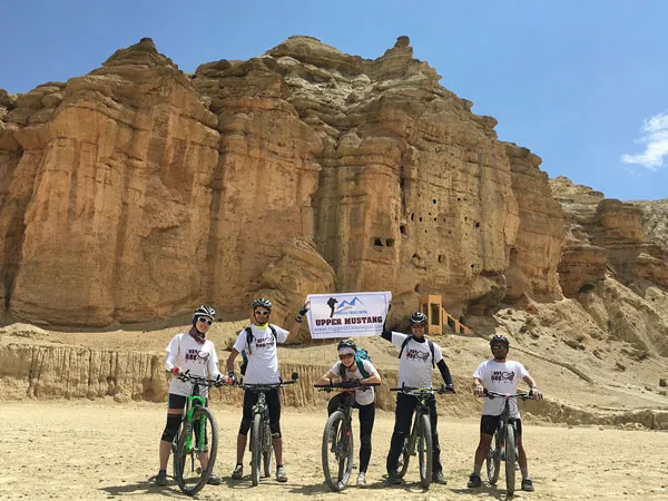 A group of mountain bikers wearing helmets, matching shirts, and sunglasses standing with their bikes holding an Upper Mustang banner in front of high, eroded sandstone cliffs