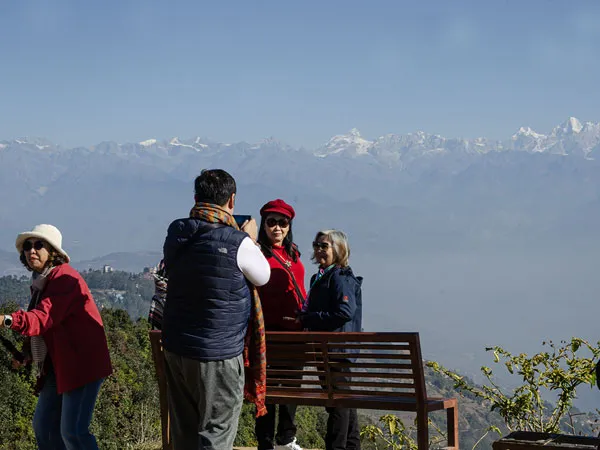 Tourists standing near a wooden bench on a hillside, taking photos of the Himalayan landscape