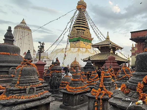 Numerous small stone votive stupas adorned with orange marigold garlands in the foreground, with the golden spire and eye-painted tower of Swayambhunath Stupa under a cloudy sky in Kathmandu