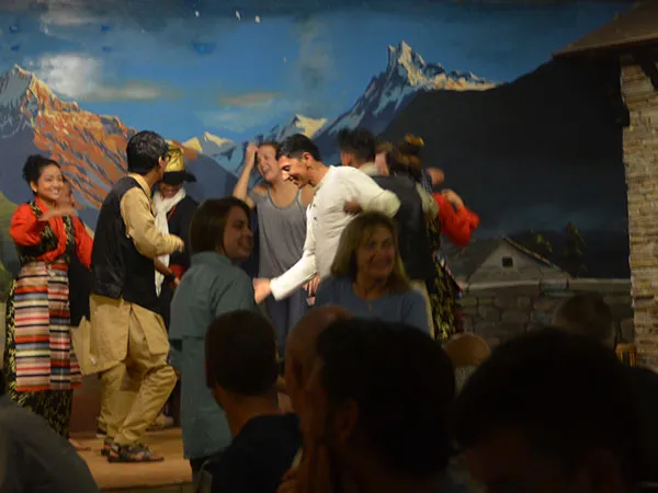 International tourists dancing enthusiastically alongside traditional Nepalese cultural performers on stage in front of a Himalayan backdrop painting.