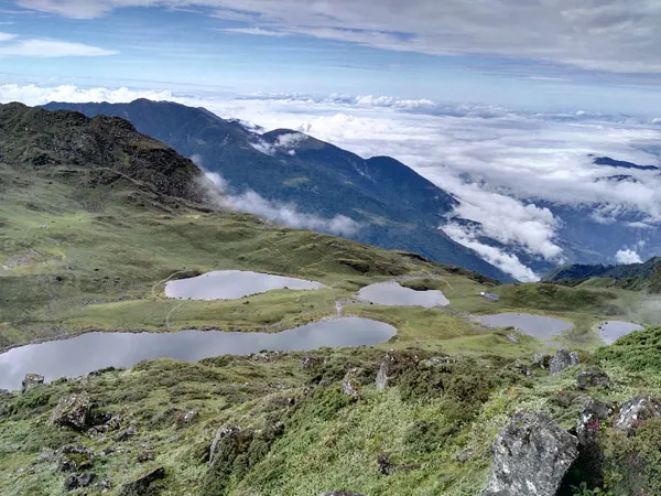 High-angle aerial landscape view of the five sacred glacial lakes of Panch Pokhari spread across green alpine pastures with surrounding mountain ridges and clouds below