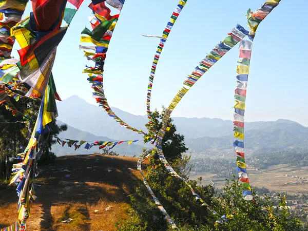 Multiple strands of colorful Tibetan Buddhist prayer flags fluttering across a hilltop path overlooking the green valley below