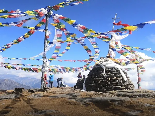 Trekkers gathered around a stone cairn wrapped in vibrant colorful Buddhist prayer flags on the rocky summit of Pikey Peak under a blue sky