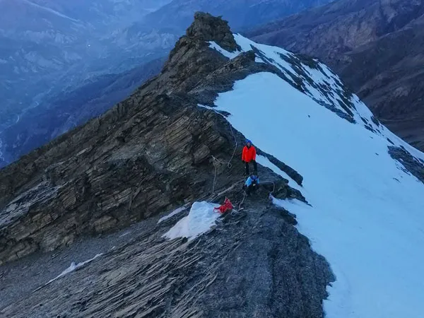 High-altitude climbers in bright red down jackets carefully navigating a narrow, snow-dusted rock ridge on Pisang Peak
