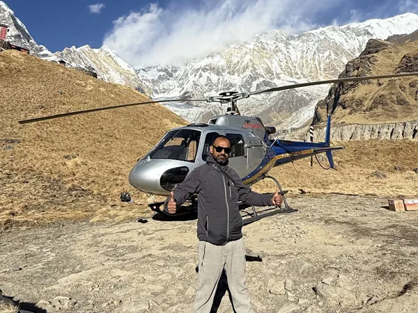 A man wearing sunglasses and a grey jacket giving a thumbs-up gesture in front of a silver and blue helicopter landed at Annapurna Base Camp, surrounded by snow-capped Himalayan peaks