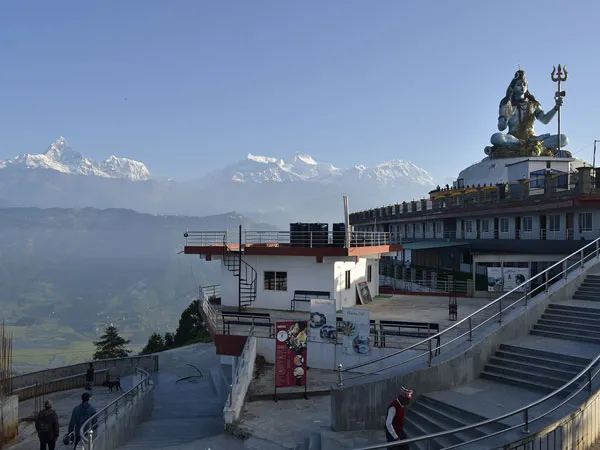 A towering seated statue of Lord Shiva holding a trident on a white dome stupa platform at Pumdikot hilltop, featuring concrete staircases, viewing terraces, and the snow-covered Annapurna Himalayan range in the background