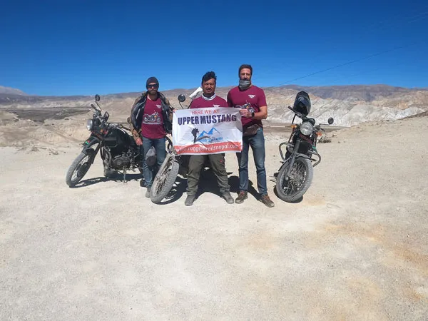 Three adventure riders standing with motorcycles on an arid dirt road in Upper Mustang, holding up a banner that reads 'Here We At Upper Mustang