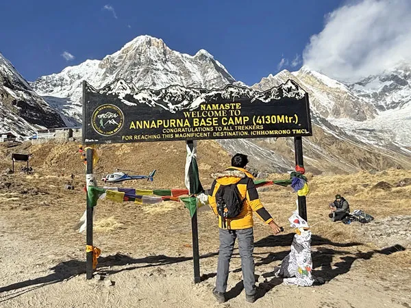 A trekker in a yellow jacket standing in front of the Annapurna Base Camp sign at 4,130 meters, with snow-capped Himalayan peaks in the background.
