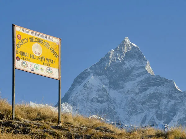 Welcome signboard at Khumai Danda (3,245m) on the Great Machhapuchhre Trail with the snow-capped Machhapuchhre (Fishtail) mountain in the background under a clear blue sky.