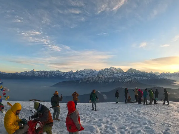group of trekkers standing on a snow-covered mountain summit with prayer flags, watching a bright sunrise over the panoramic Everest and Himalayan range