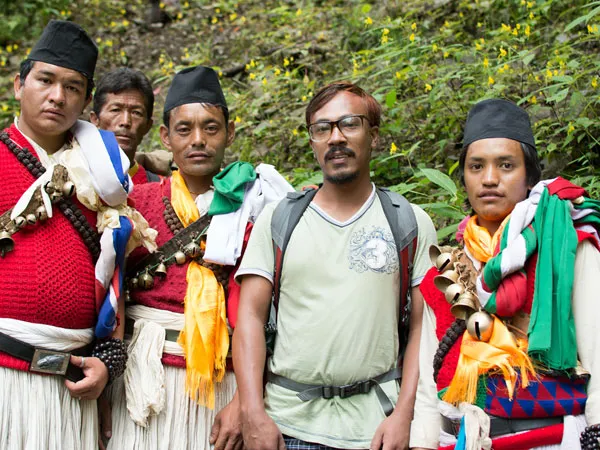 A trekker standing alongside traditional Tamang shamans (Dhami/Jhakri) wearing ritual attire, caps, and brass bells during the Tamang Heritage Trek in Nepal.