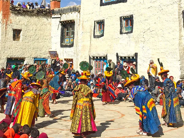 Buddhist monks wearing ornate brocade robes and traditional hats performing sacred ritual dances in the open courtyard of the walled city of Lo Manthang, surrounded by crowds of locals and tourists