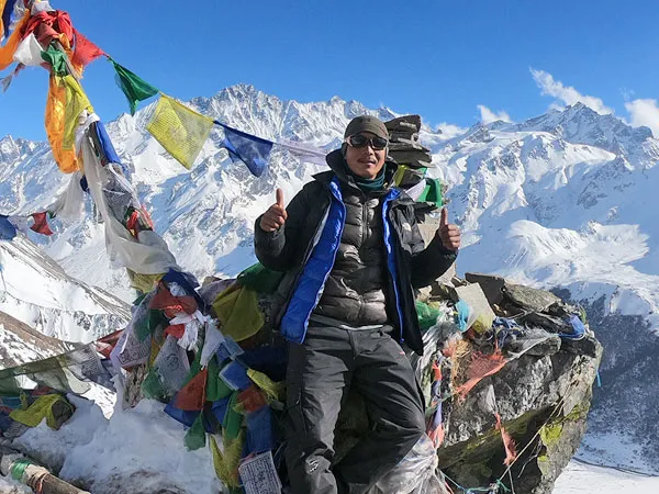 A trekker wearing warm winter gear giving a thumbs-up at a high mountain viewpoint surrounded by prayer flags and snow-capped Himalayan peaks on the Langtang Valley Short Trek.