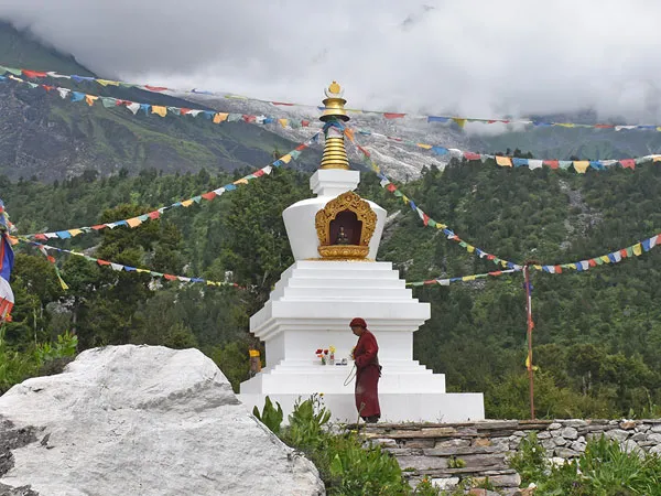 A Buddhist monk praying at a white chorten decorated with prayer flags against a lush mountain backdrop in Tsum Valley