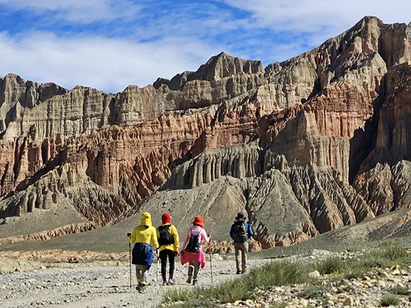 Four trekkers with backpacks and hiking poles walking along a dirt trail against the backdrop of massive, stratified red rock cliffs and canyons under a clear blue sky