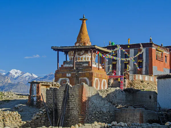 Traditional Buddhist stupa and monastery with colorful prayer flags in Upper Mustang, Nepal, against a clear blue sky and distant snow-capped mountains.