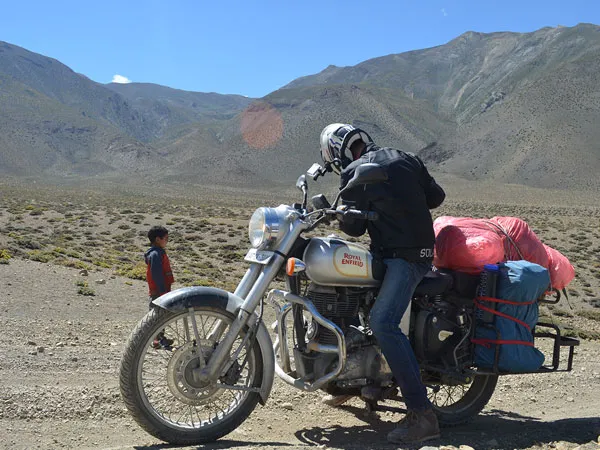 A motorcyclist wearing a helmet and riding gear seated on a classic silver Royal Enfield loaded with luggage along a dirt road in a dry, mountainous landscape