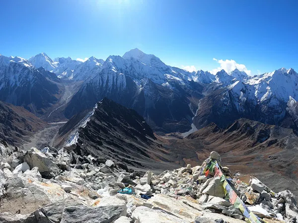 A high-altitude mountain summit adorned with colorful Buddhist prayer flags, overlooking rugged rock ridges and snow-capped Himalayan ranges under a clear blue sky