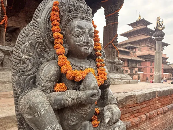 A detailed close-up of an ancient black stone sculpture of a guardian deity adorned with vibrant orange marigold garlands, with traditional multi-roofed Newari temples of Patan Durbar Square in the background