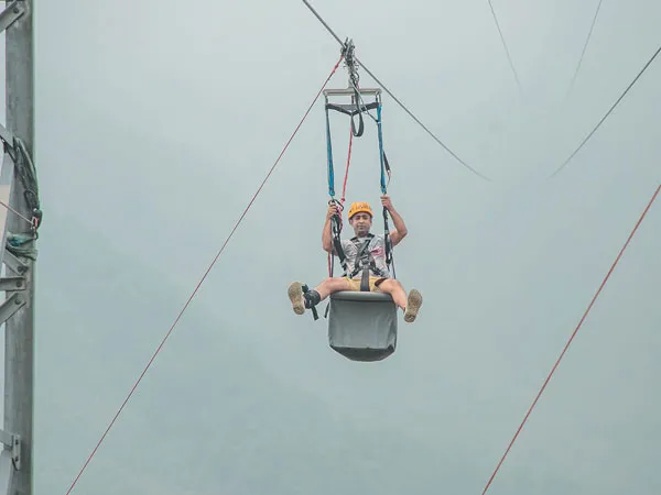A person strapped into a harness helmet sitting in mid-air on the ZipFlyer zipline system against a misty mountain backdrop