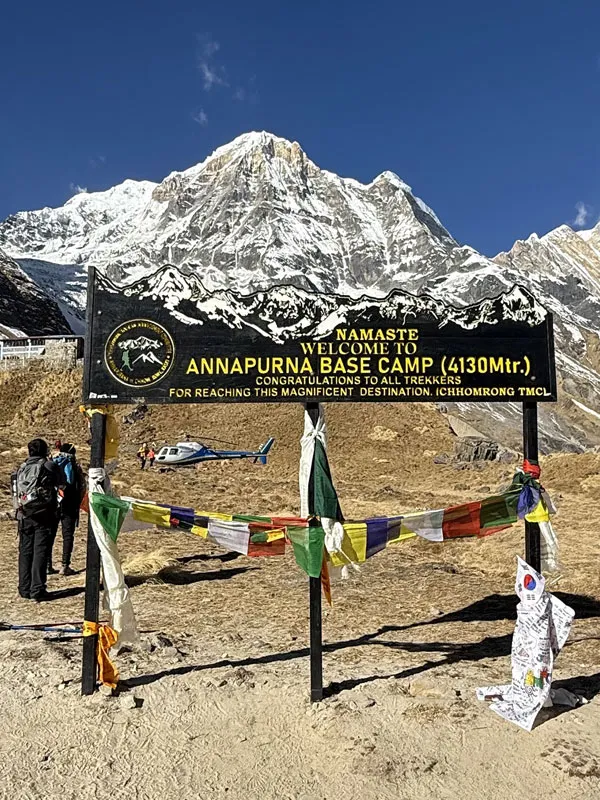 Trekkers standing near the black Welcome to Annapurna Base Camp 4130m signboard decorated with prayer flags, with snow-capped Annapurna peaks and a helicopter in the background