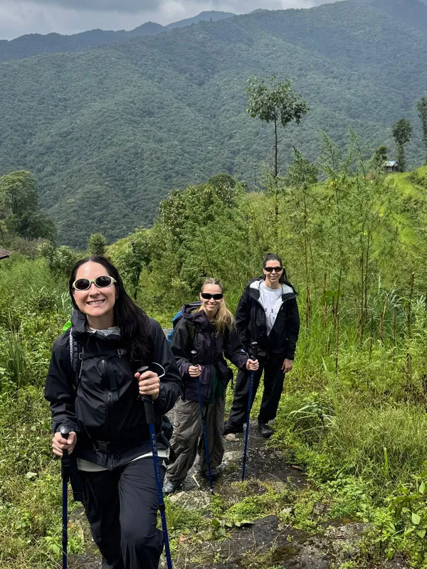 Three female trekkers with backpacks and trekking poles smiling while walking along a lush green trail on the Helambu Circuit with forested hills in the background