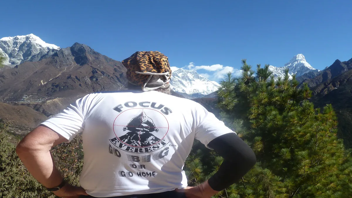 A trekker wearing a graphic t-shirt reading Focus Everest standing with hands on hips, looking out towards snow-covered Himalayan peaks and Ama Dablam