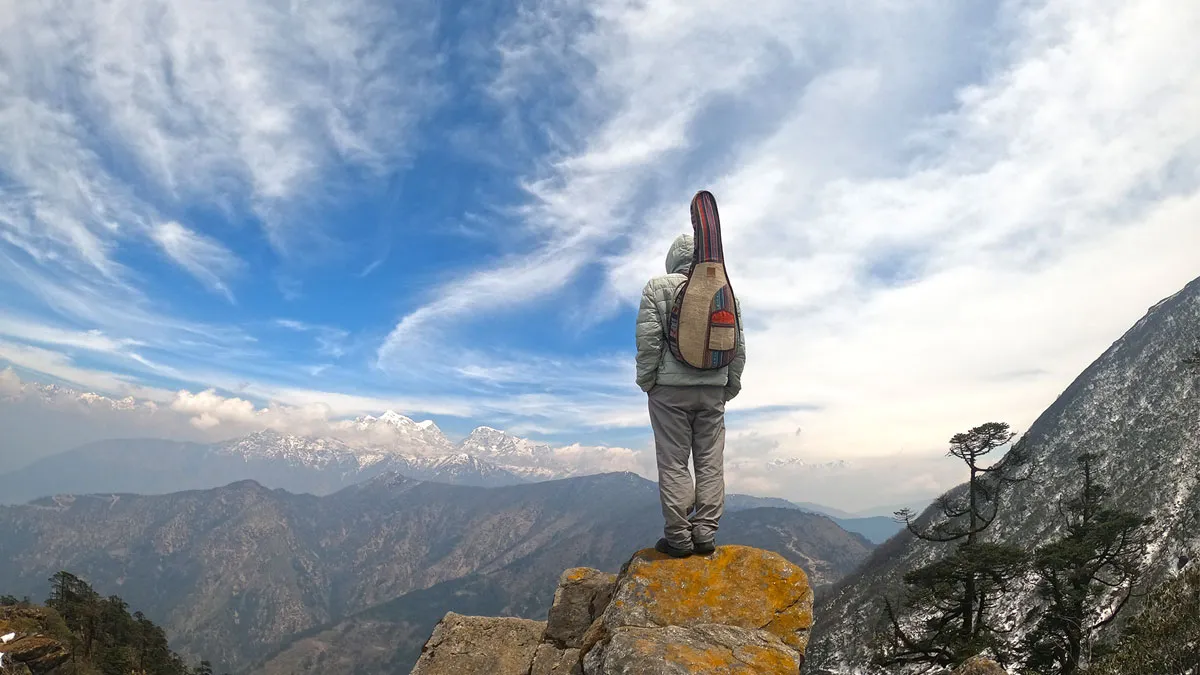 A trekker carrying an instrument case standing atop a rocky outcrop looking out at panoramic mountain valleys and snow-capped Himalayan peaks under a bright cloudy sky during the Pikey Peak Trek